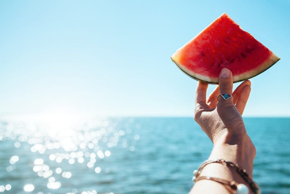A hand holding a slice of watermelon against a sparkling water backdrop, symbolizing summer at City Base Vista in San Antonio, Texas.